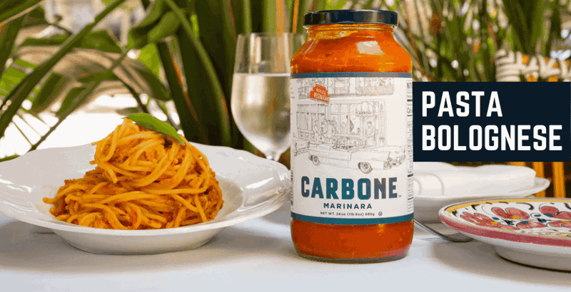 A plate of Carbone Pasta Bolognese next to a jar of Carbone Marinara, a wine glass in the back. 